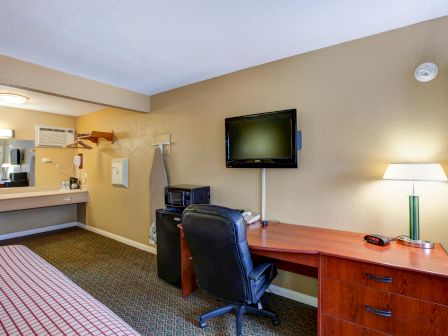 A hotel room features a bed, desk with chair, TV, lamp, microwave, and an open sink area. The walls are beige and the carpet is dark.