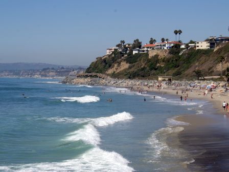 A scenic beach with waves rolling in, people swimming and relaxing, and houses perched on a cliff in the background, under a clear blue sky.