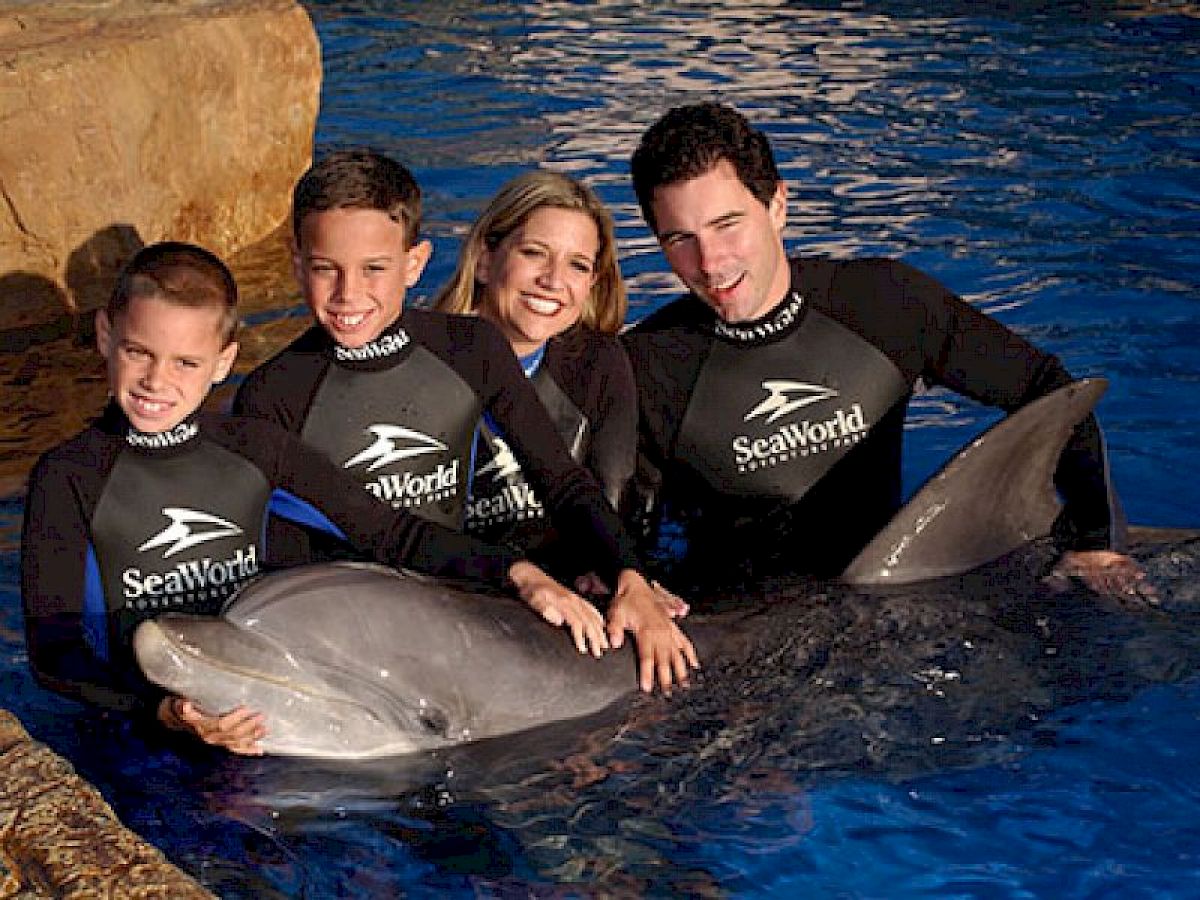 A family wearing wetsuits and posing with a dolphin in a water setting at SeaWorld, all smiling and closely gathered around the dolphin.