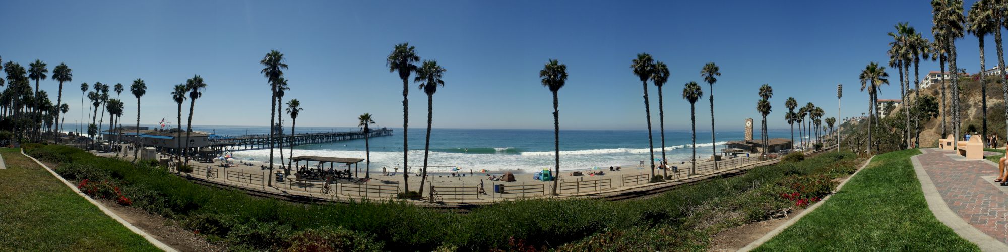A beachfront scene with palm trees, a pier extending into the ocean, and people enjoying the sandy beach and waves under a clear blue sky.