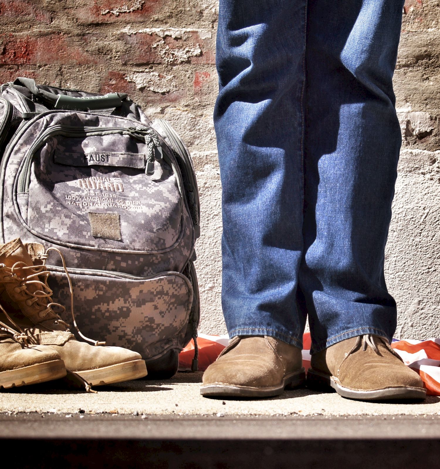 A person in jeans stands beside military boots, a camouflage backpack, and a partially visible flag on the ground, against a brick wall.