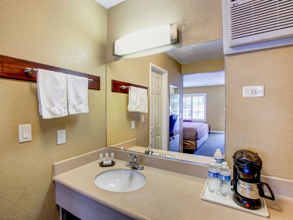 A bathroom vanity area with a sink, mirror, towel rack, toiletries, water bottles, and a coffee maker, adjacent to a room with a bed seen through the door.