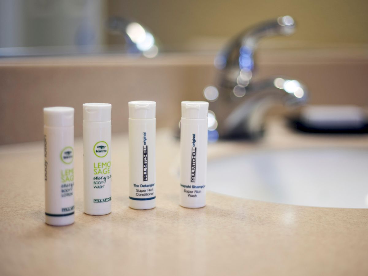 The image shows four small toiletry bottles placed on a bathroom countertop near a sink faucet.