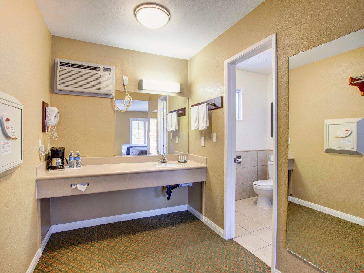 A hotel room's washroom with a large vanity, mirror, toiletries, towels, wall-mounted hairdryer, thermostat, and a separate toilet area.