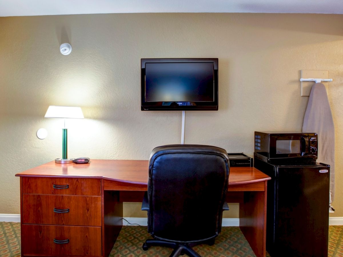 The image shows a desk with a chair, a wall-mounted TV above the desk, a desk lamp, a mini-fridge, a microwave, and a coat hanger in a room.