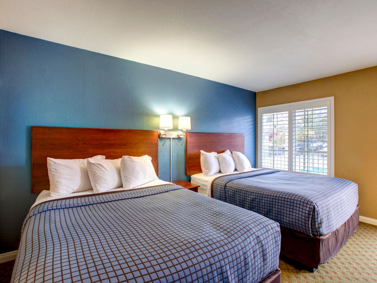 The image shows a hotel room with two double beds, each with four pillows, modern decor, a blue accent wall, and a window with plantation shutters.