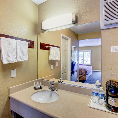 A hotel room's vanity area with a sink, towels, a light fixture, bottled water, a coffee maker, and a view into the bedroom.