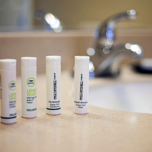 The image shows four white toiletries containers on a bathroom countertop near a sink, with a blurred faucet in the background.