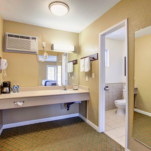This image shows a hotel room with a vanity area, mirror, towels, and a toilet visible through an open door.