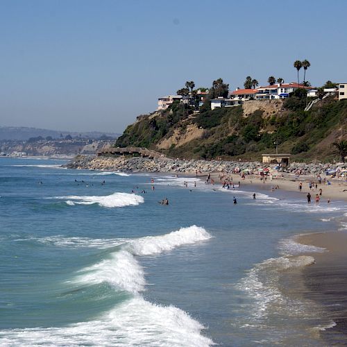 A scenic beach with waves crashing on the shore, people swimming and relaxing, and houses on a hillside overlooking the ocean, ending the sentence.