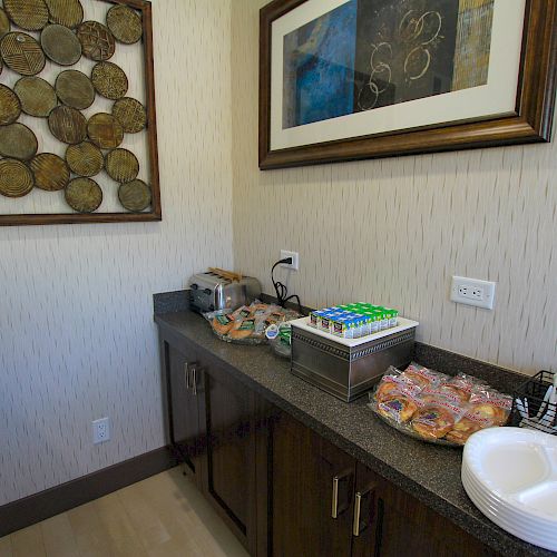 A small counter setting with snacks, plates, utensils, a toaster, and artwork on the wall. There appears to be a variety of packaged snacks and cereals.
