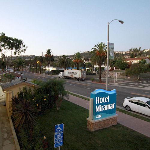 The image shows the exterior view of Hotel Miramar situated along a busy road with palm trees and parked vehicles at dusk.