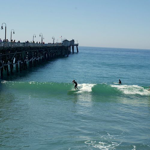 The image shows a pier extending into the ocean, with surfers riding waves near it. The sky is clear and the water is blue.