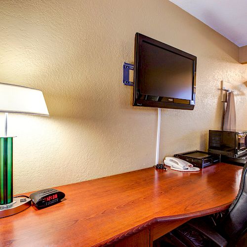 The image shows a hotel room desk setup with a lamp, clock, TV, microwave, and coffee maker, along with a chair and some hangers in the background.
