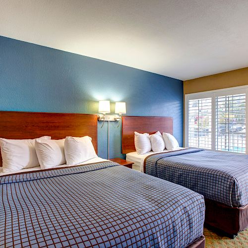 This image shows a hotel room with two large beds, each with four pillows, a blue accent wall, a window with shutters, and a framed picture on the wall.