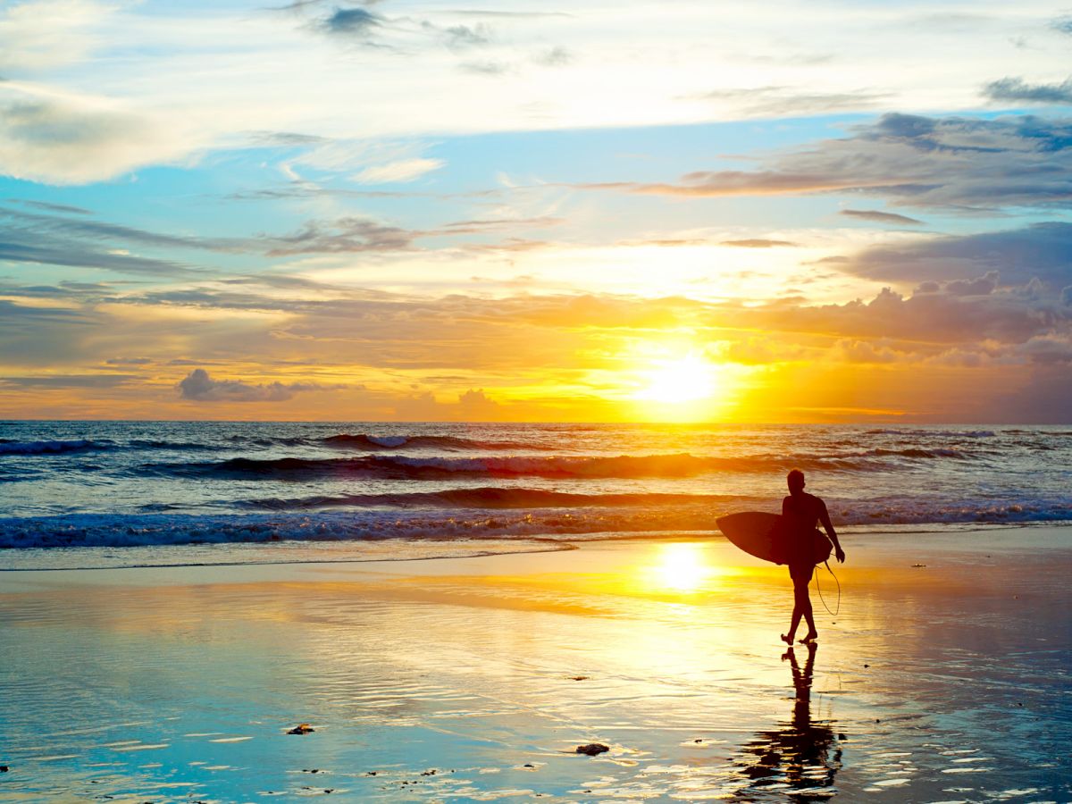 A person with a surfboard walks along a beach at sunset, with vibrant colors reflecting off the wet sand and waves in the background.