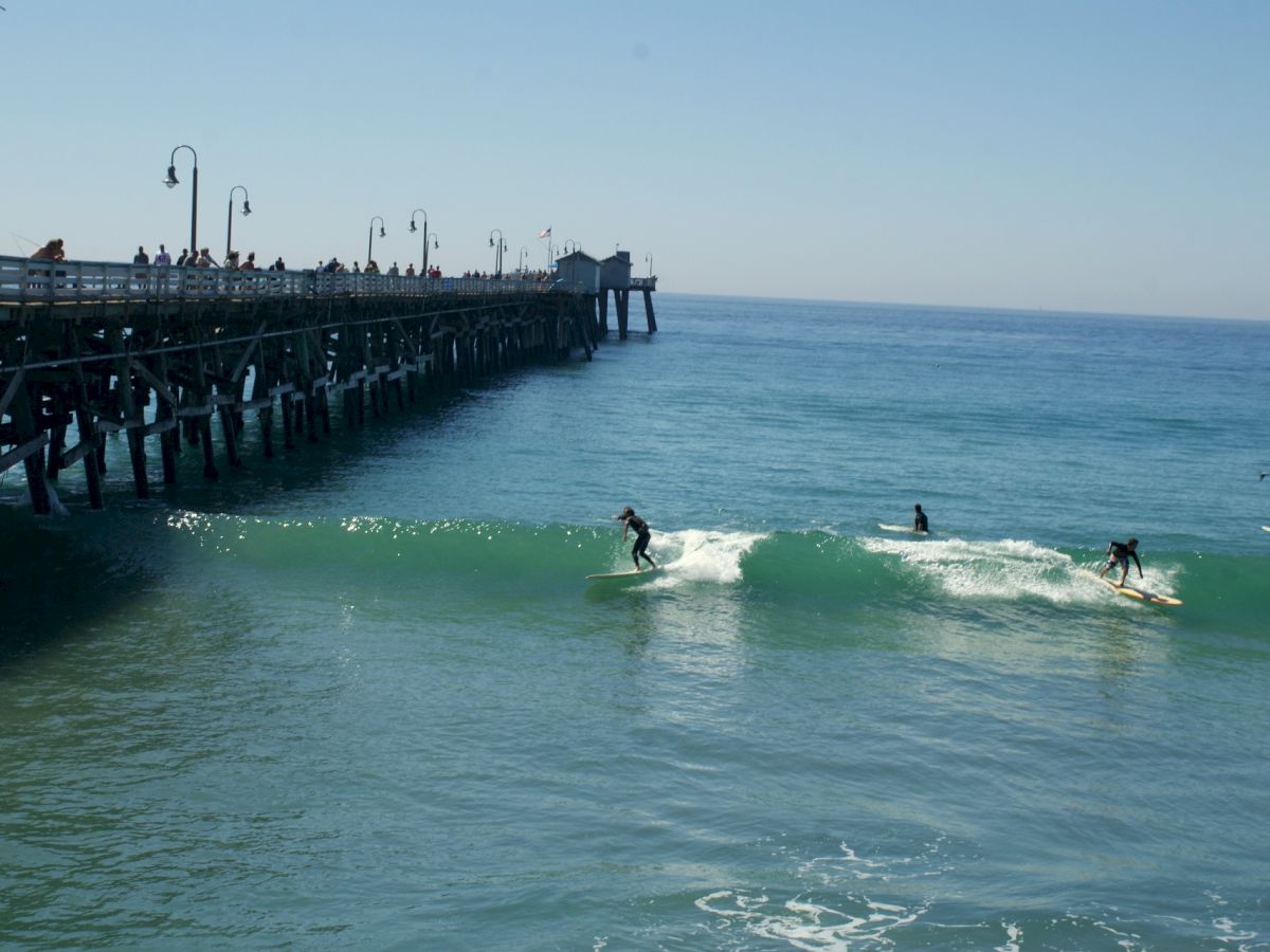 Surfers ride waves near a pier with people observing from above on a clear, sunny day.