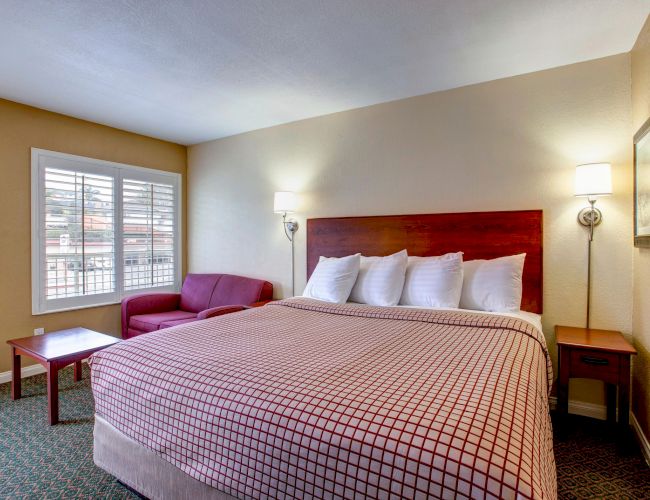 A cozy hotel room with a king-sized bed, decorative pillows, two lamps, a red sofa, a coffee table, and a window with shutters.